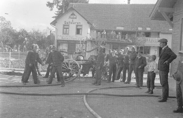 Zu sehen ist ein altes Foto, auf dem die Feuerwehr bei einer Übung auf dem damaligen Gemeindehausplatz um 1910 zu sehen ist.